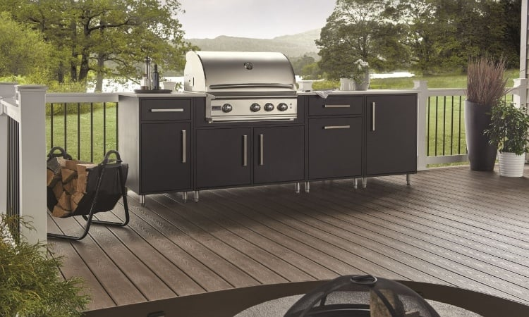 Modern outdoor kitchen on a spacious deck featuring a stainless steel built-in grill, dark storage cabinets, fire pit and scenic backyard views.
